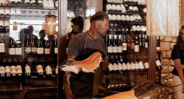 Een man staat in een restaurant met een gesneden vis en wijnflessen op de achtergrond.