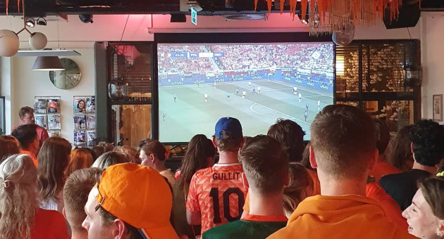 Een groep mensen in oranje kleding kijkt naar een voetbalwedstrijd op een groot scherm.