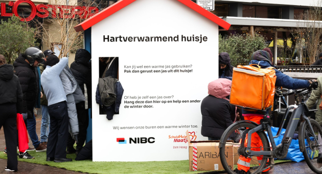 Houten huisje op de Grote Markt in Den Haag waar mensen omheen staan waaraan warme jassen worden uitgedeeld. 