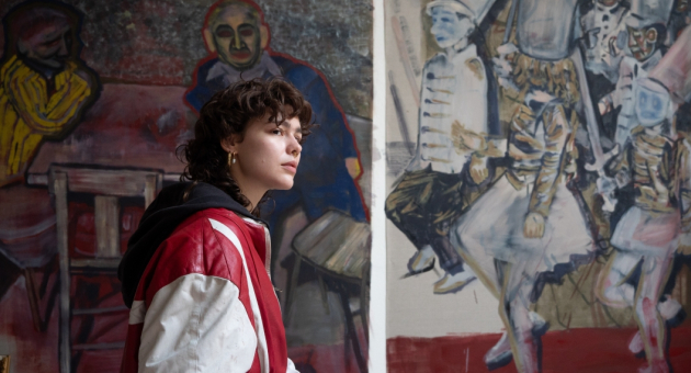 Een jonge vrouw met krullend haar staand voor schilderijen in een galerie.