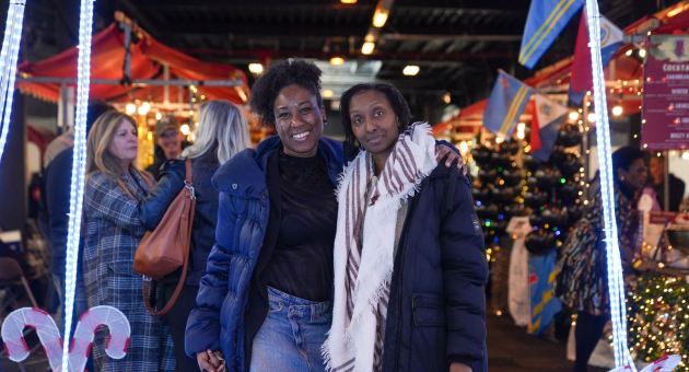 Twee vrouwen poseren samen op een kerstmarkt, omringd door kraampjes en feestelijke verlichting.