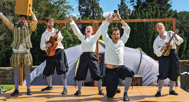 Six actors in historical costumes perform joyfully on a stage.