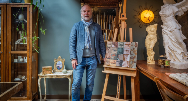 Een man staat in een atelier naast een schildersezel met gezichten.