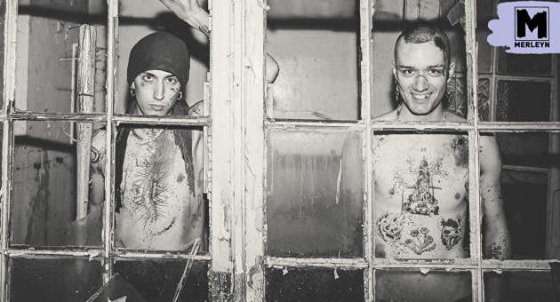 Twee mannen met tattoos staan achter een vergrendeld raam.