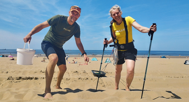 Een man en vrouw staan op het strand, hij draagt een T-shirt en shorts, zij een gele shirt en sportkleding.