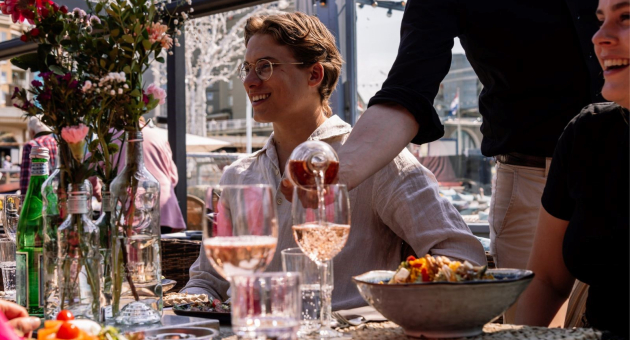 Mensen genieten van een maaltijd op een zonnig terras met bloemen en drankjes.