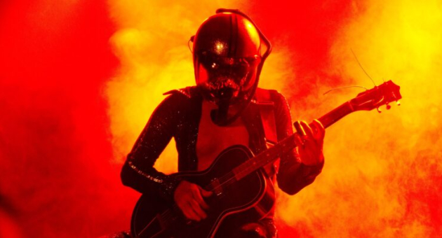 Een muzikant in een ruimtehelm speelt gitaar tegen een achtergrond van felgekleurde rook.