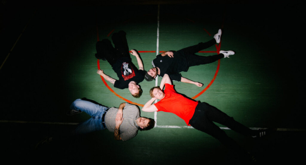 Vijf jongeren liggen op een sportveld in een cirkel.