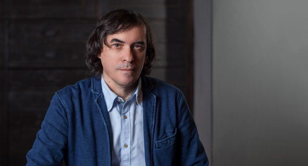 A man with dark, medium-length hair wearing a blue jacket.