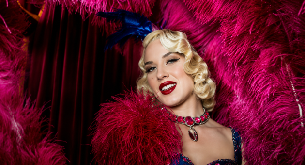 Magical Burlesque By AMI. Een vrouw met blond krullend haar in een opvallende, glinsterende jurk met een roze veren boa.