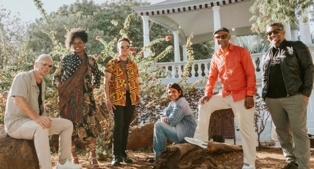 Zes mensen van de muziekgroep CaboCubaJazz poseren in een tuin met bloemen.