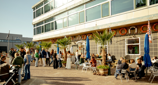 Mensen op een zonnig terras bij het Zeemanshuis in de haven van Scheveningen, omringd door palmbomen en maritieme details  @charlottevdgaag