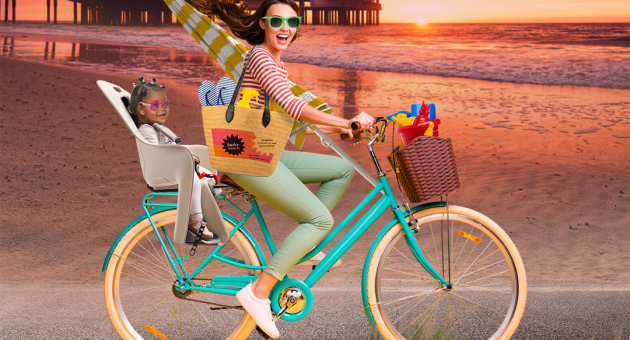 Een vrouw fietst met een kind en strandspullen, op de achtergrond de zee en een reuzenrad.