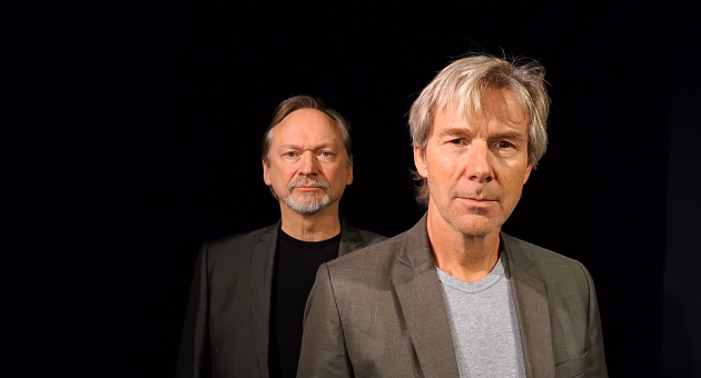 Twee mannen met grijze jassen staan in een donkere ruimte