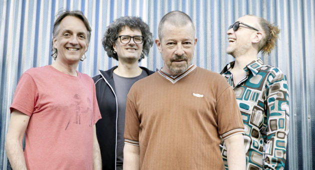 Vier mannen poseren lachend voor een metalen achtergrond.