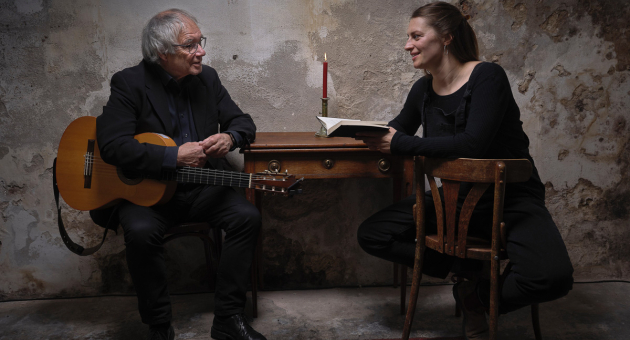 Een man met een gitaar en een vrouw met een boek zitten aan een tafel.