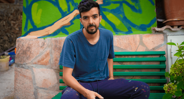 Een man zit op een groene bank met een borstel in zijn hand, voor een muurschildering.