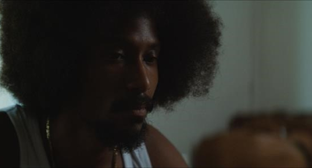 Filmfragment  van een man met afro haar kijkt serieus naar links. Srefidensi Dey – 50 jaar onafhankelijkheid van Suriname