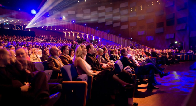 A packed theater audience watching the stage.