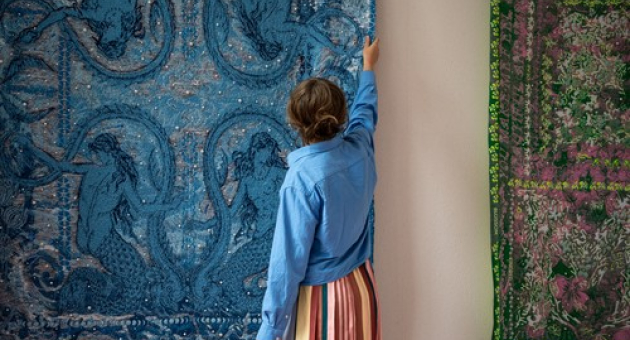 Een vrouw in een blauwe blouse staat voor twee wandtapijten. Foto: Studio Dewi, 2026