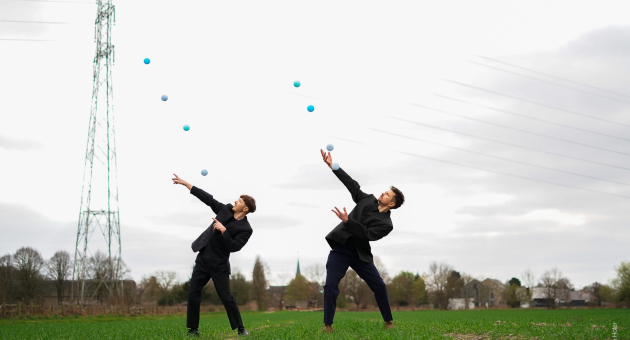 Jam.Shenanigans – Splash. Two young men in black suits juggling balls in a green field.