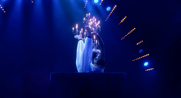 Een performer in een donkerblauwe setting met een kroonluchter, elegant in beweging.
