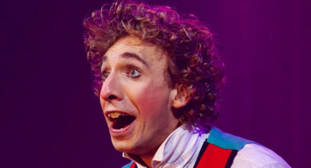 Een jonge man met krullend haar glimlacht breed, gekleed in een kleurrijk kostuum.