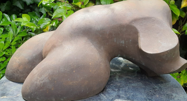 Kim Kroes- Ode aan de vrouw. Abstract bronzen sculptuur met ronde vormen, omringd door groene bladeren.