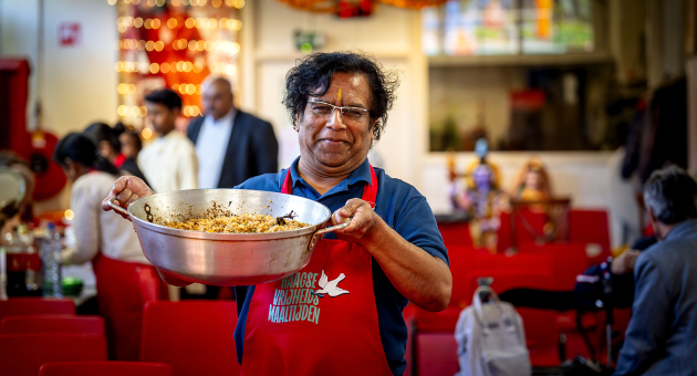 Een man in een rood schort houdt een pan met gekruid rijstgerecht vast in een sfeervolle, drukke ruimte.