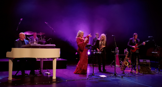 Zangeres in rode glitterjurk met liveband op podium, paarse belichting en diverse instrumenten zichtbaar