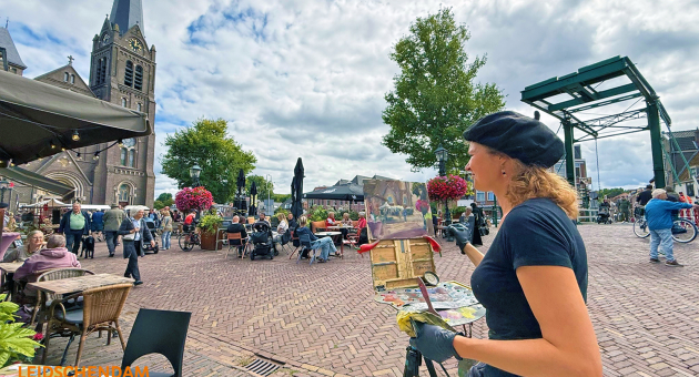 Een schilderes met een palet schildert op een plein, omringd door mensen.