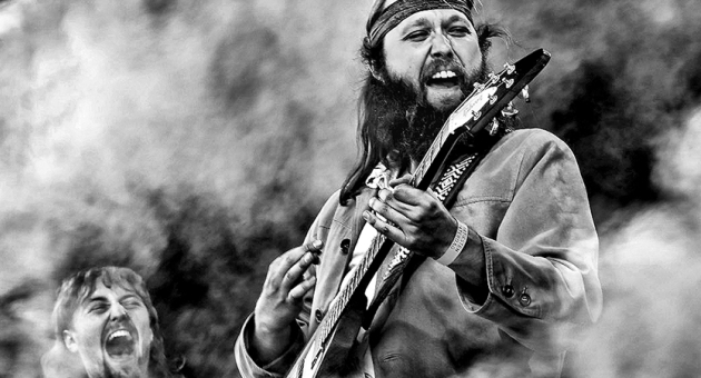Een rockmuzikant speelt gitaar met veel passie, terwijl een andere muzikant op de achtergrond enthousiast meezingt.