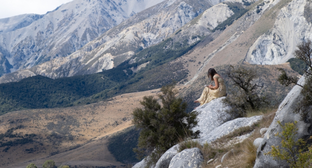 Een vrouw in een gouden jurk zit op een rots met bergen op de achtergrond.