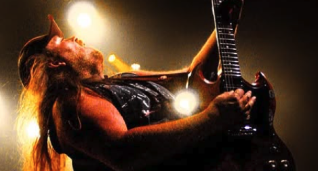 Een man met lang haar speelt electrische gitaar op een podium met lichteffecten.