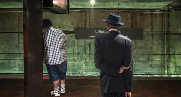 Twee mannen wachten op een metro bij een station, de één in casual kleding en de ander in een pak met een hoed.