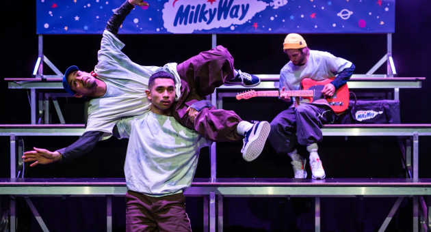 Three dancers performing on stage with a guitar and MilkyWay branding.