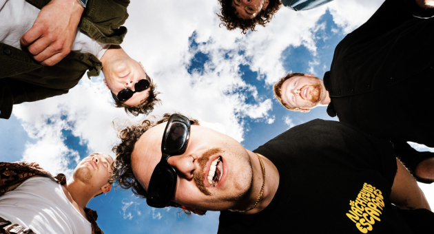 Vijf vrienden in een cirkel, kijkend naar de camera met een blauwe lucht op de achtergrond.