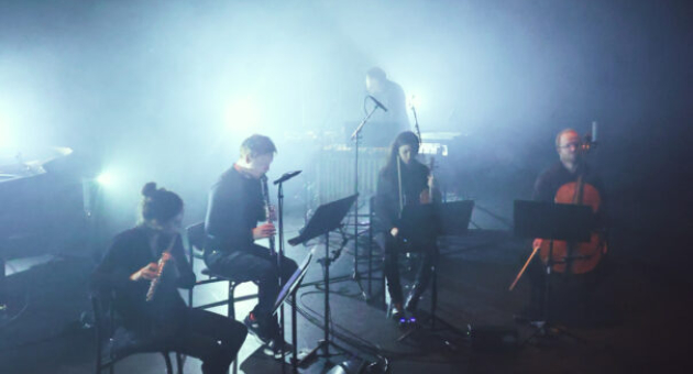 Zes muzikanten spelen samengevoegd in een mistige omgeving met blauwe lichten.