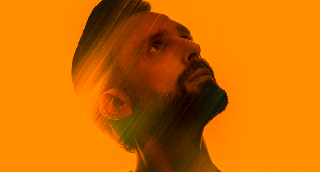 Portret van zanger Nick Schilder schuin omhoog kijkend blendend in de oranje achtergrrond.