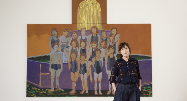 Kunstenaar poseert voor een groot schilderij van een groep kinderen in gestreepte badpakken, met een felgele fontein boven het werk. De foto is genomen in een moderne tentoonstellingsruimte.