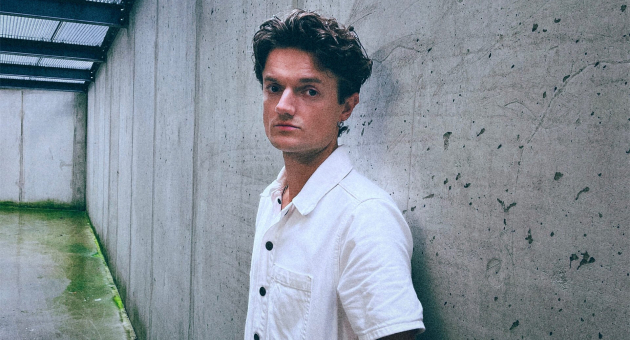 Zanger Chet met gekruld haar staat tegen een betonwand.