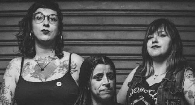 Drie vrouwen met tatoeages, poseren trots voor een muur.