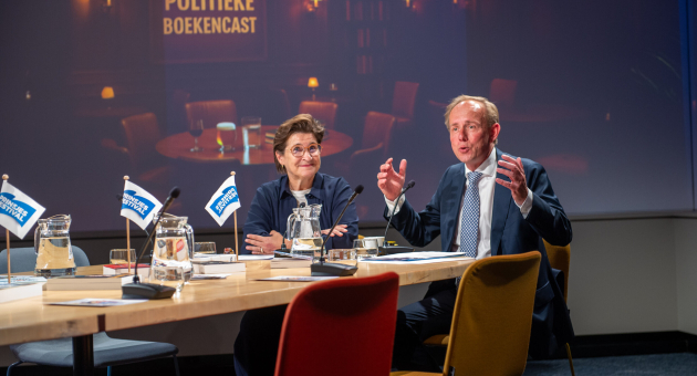 Twee sprekers discussiëren aan een tafel met boeken en glazen.