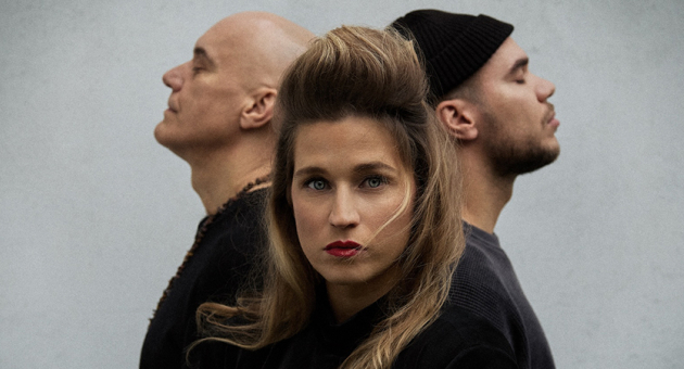 Drie personen met opvallende poses. Een vrouw met lange haren en rode lippen, met twee mannen in de achtergrond. De mannen zijn met hun rug naar de camera gekeerd.