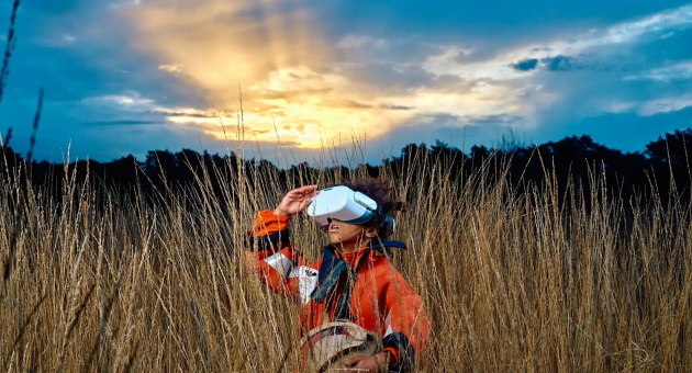 Installatie: ERGENS (8+). Een persoon met een VR-bril staat in hoog gras en kijkt naar de lucht tijdens zonsondergang.