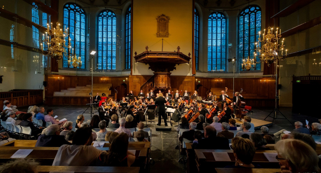 Een orkest leidt een uitvoering in een sfeervolle zaal met grote ramen.