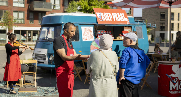 Een man in een schort praat met twee bezoekers op een markt.