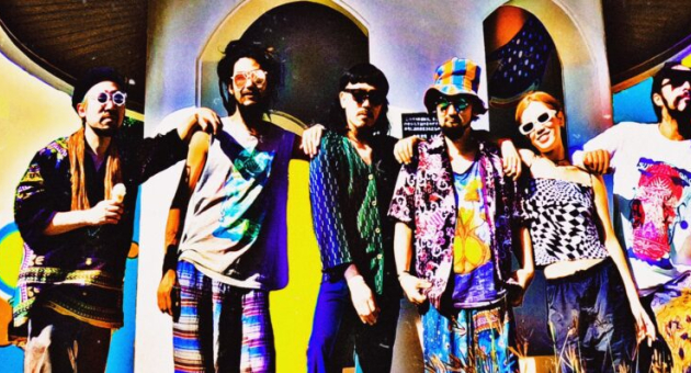 De bandleden van Super Jet Kinoko poseren in een holografische setting, omringd door lichtprojecties en transparante kleurreflecties. De compositie geeft een futuristisch en vervreemdend effect, passend bij hun trance-achtige stijl.