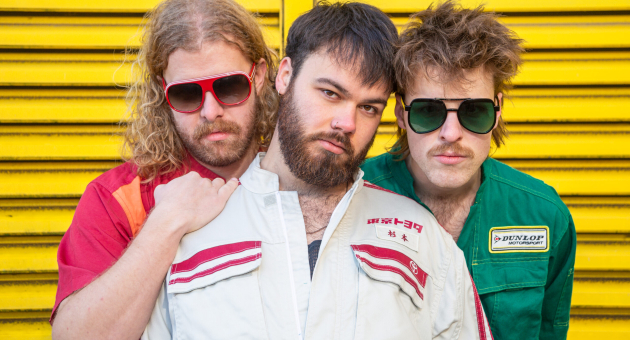 Drie mannen van de band TaxiTaxi poseren in kleurrijke kleding tegen een gele achtergrond.