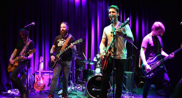 Vijf muzikanten op een podium met lichten, spelen live muziek.